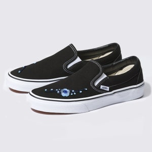 Puede incluir: Zapatillas Vans negras de slip-on con suelas blancas y bordado floral azul en el lateral. El logotipo de Vans es visible en el lateral de la zapatilla.