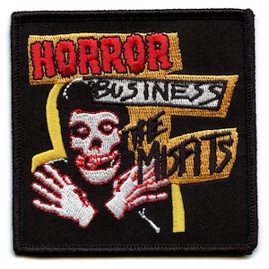 May include: A black embroidered patch with a yellow and white sign that reads "Horror Business The Misfits". A cartoon skull with red eyes and a red mouth is in the center of the patch.