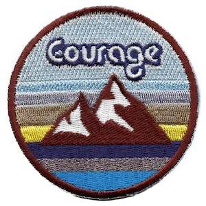 Op de afbeelding: Een ronde geborduurde patch met een blauwe, witte en bruine berg scène. Het woord "Courage" is in blauw boven de bergen geborduurd.