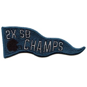 Peut inclure: Un fanion bleu en feutre avec du texte brodé en blanc qui dit "2X SB CHAMPS". Une silhouette noire d'un casque de football se trouve à gauche du texte.