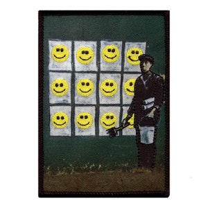 May include: Embroidered patch featuring a street artist painting yellow smiley faces on a green wall. The artist is depicted in black clothing, holding a brush and bucket. The patch has a black border.