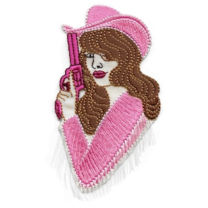 May include: Embroidered patch of a woman wearing a pink cowboy hat and holding a pink gun. The patch has a fringed edge.