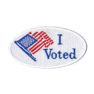 May include: White oval sticker with the text "I Voted" in blue embroidery. A red, white, and blue American flag is embroidered on the left side of the sticker.