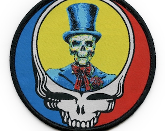 May include: Round patch featuring a stylized skull wearing a blue top hat and suit with a red bow tie and rose. The design is set against a yellow background, framed by blue and red sections, and a black border.