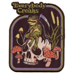 May include: A green frog wearing a black hooded robe sits on a white skull with a scythe in its hand. The skull is surrounded by red and yellow mushrooms. The background is dark brown with the text "Everybody Croaks" in a yellow and brown font.