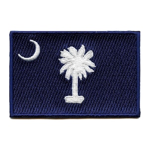May include: A blue embroidered patch featuring the South Carolina state flag. The flag depicts a white palmetto tree and a crescent moon on a dark blue background.