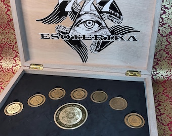 Puede incluir: Caja de madera abierta con la palabra "ESOTERIKA" y un símbolo de ojo. Contiene varias monedas doradas con símbolos y texto. La caja está sobre una superficie estampada.