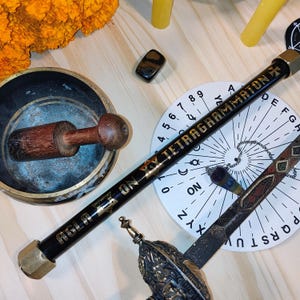 May include: A black wooden wand with gold lettering that reads "AGLA ON TETRAGRAMMATON", resting on a white circular divination tool with a pendulum. The wand is surrounded by other divination tools, including a wooden bowl, a crystal, and a candle.