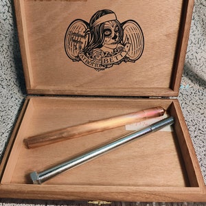 May include: A wooden box with a hinged lid. The lid has a black and white illustration of a woman with wings and the text "Deadwood Tattoo Co. Fat Bottom Betty". The box is open and contains a copper pipe and a metal rod.