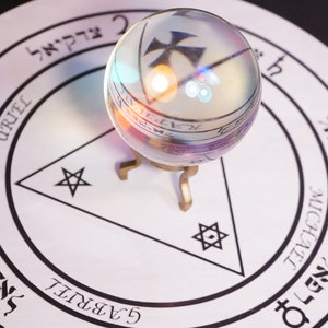 Op de afbeelding: Een kristallen bol staat op een gouden standaard bovenop een witte cirkel met zwarte markeringen en Hebreeuwse tekst. De cirkel bevat een driehoek met een vijfpuntige ster op elk punt.