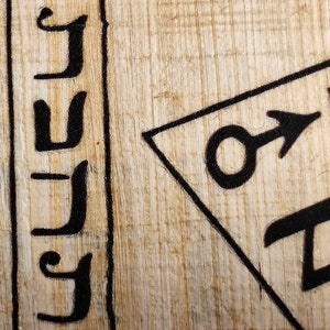 Op de afbeelding: Close-up van een gedeelte van papyrus met zwarte inktmarkeringen. De afbeelding toont gestileerde zwarte letters en een geometrisch symbool met een pijl. De papyrus heeft een lichtbeige kleur met zichtbare houtnerf.