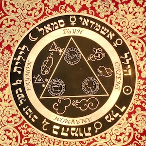 May include: A circular, gold-colored talisman with intricate symbols and text in a foreign language. The center features a black triangle with additional symbols. The background is a red and gold patterned fabric.