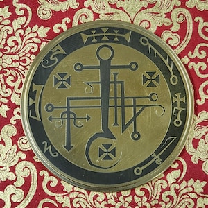 Może przedstawiać: Okrągły, mosiężny metalowy przedmiot z czarną obwódką. Zawiera skomplikowane geometryczne wzory i symbole, w tym krzyże, strzałki i linie. Tło to czerwona tkanina ze złotym wzorem kwiatowym.