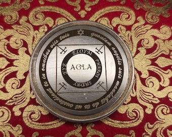 Puede incluir: Un colgante circular plateado con texto y símbolos grabados. El diseño central presenta la palabra "AGLA" dentro de un círculo, rodeada de otras inscripciones y formas geométricas. El colgante descansa sobre una tela estampada roja y dorada.