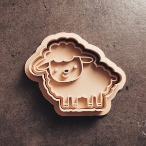 May include: A beige plastic cookie cutter shaped like a sheep. The sheep has a simple face with two eyes, a nose, and a mouth.
