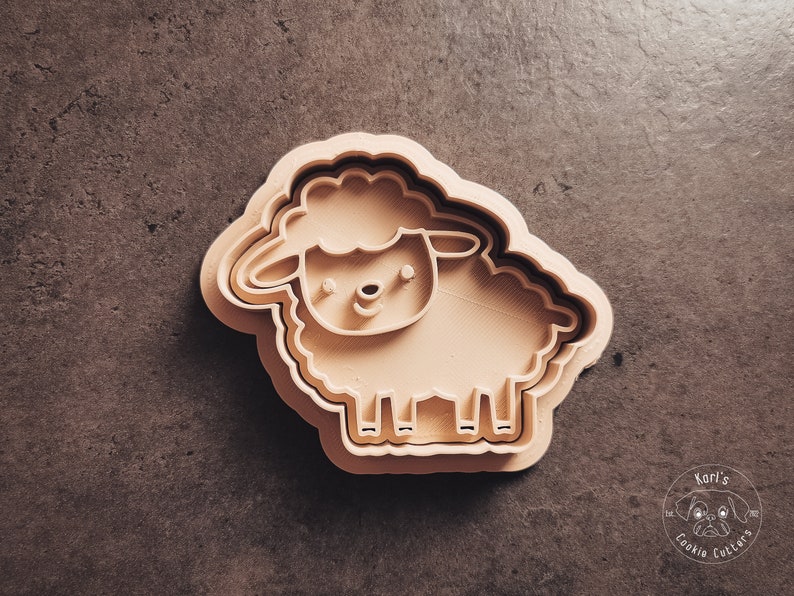 May include: A beige plastic cookie cutter shaped like a sheep. The sheep has a simple face with two eyes, a nose, and a mouth.