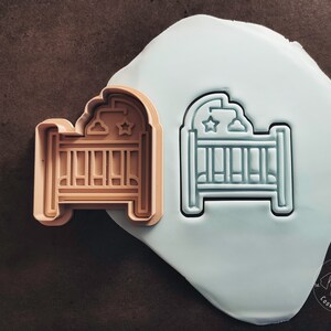 Könnte beinhalten: Ein brauner Plastik-Ausstecher in Form eines Babybettes mit einem Stern- und Wolkenmuster. Der Ausstecher wird neben einer weißen Plastikform desselben Designs gezeigt.