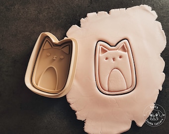 Puede incluir: Primer plano de una masa de galletas rosa con un cortador de galletas con forma de gato presionado en ella. El cortador de galletas es beige y tiene un diseño de cara de gato. El texto "Lori's Cookie Cutters" es visible en la esquina inferior derecha de la imagen.