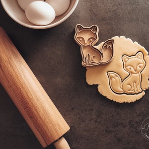 Puede incluir: Primer plano de dos cortadores de galletas con forma de zorro sobre una superficie marr&oacute;n. Los cortadores est&aacute;n hechos de metal y tienen un acabado marr&oacute;n. Los cortadores est&aacute;n junto a un rodillo y un taz&oacute;n de huevos. El texto "Kori's Cookie Cutters" es visible en la esquina inferior derecha de la imagen.