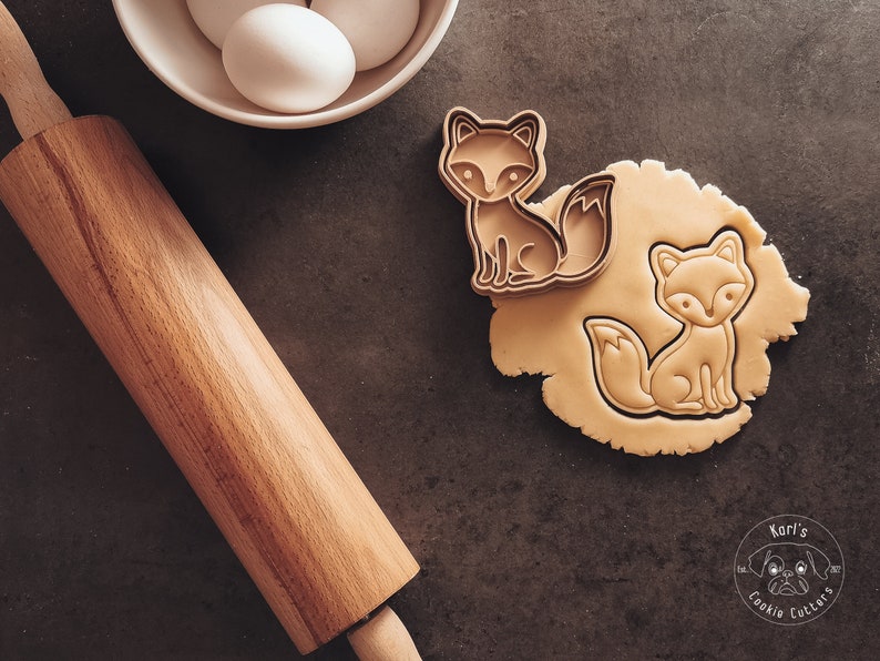 Puede incluir: Primer plano de dos cortadores de galletas con forma de zorro sobre una superficie marr&oacute;n. Los cortadores est&aacute;n hechos de metal y tienen un acabado marr&oacute;n. Los cortadores est&aacute;n junto a un rodillo y un taz&oacute;n de huevos. El texto "Kori's Cookie Cutters" es visible en la esquina inferior derecha de la imagen.