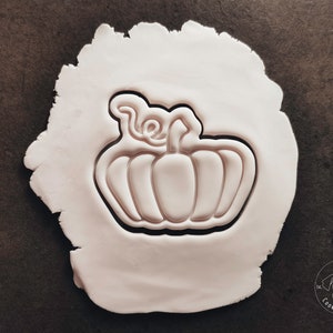 Puede incluir: Una superficie de arcilla blanca con un cortador de galletas con forma de calabaza. El cortador tiene un dise&ntilde;o detallado con un tallo y hojas.