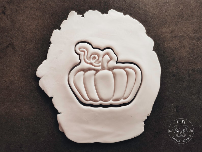 Puede incluir: Una superficie de arcilla blanca con un cortador de galletas con forma de calabaza. El cortador tiene un dise&ntilde;o detallado con un tallo y hojas.
