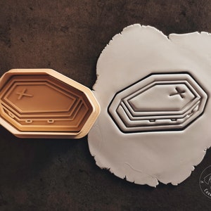 Könnte beinhalten: Ein brauner Plastik-Ausstecher in Form eines Sarges mit einem Kreuz oben drauf. Der Ausstecher wird neben einem weißen Stück Modelliermasse gezeigt, aus dem die gleiche Form ausgeschnitten ist.