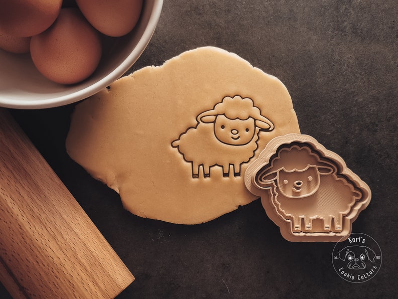 May include: A brown cookie cutter in the shape of a cartoon sheep with a smiling face. The cutter is on a brown surface next to a piece of cookie dough. The text "Kari's Cookie Cutters" is visible in the lower right corner of the image.