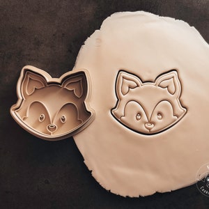 Könnte beinhalten: Ein brauner Plastik-Ausstecher in Form eines Fuchsgesichts. Der Ausstecher liegt auf einem Blatt weißen Fondants. Der Fondant hat den Abdruck des Fuchs-Ausstechers.