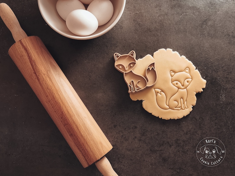 Puede incluir: Dos cortadores de galletas con forma de zorro sobre una superficie marr&oacute;n. Un cortador est&aacute; encima de una masa, el otro est&aacute; al lado. Un rodillo de madera y un taz&oacute;n de huevos est&aacute;n en el fondo. La imagen incluye el texto "Karl's Cookie Cutters".