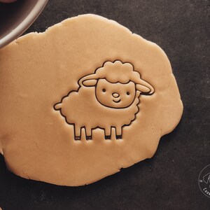 May include: A close-up of a brown cookie dough with a sheep cookie cutter pressed into it. The cookie cutter is a simple outline of a sheep with a curly head and four legs.