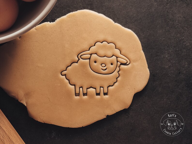 May include: A close-up of a brown cookie dough with a sheep cookie cutter pressed into it. The cookie cutter is a simple outline of a sheep with a curly head and four legs.