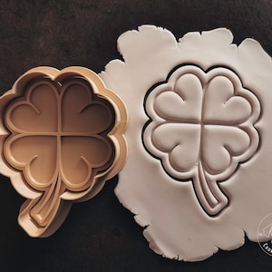 Könnte beinhalten: Ein brauner, vierblättriger Kleeblatt-Ausstecher aus Kunststoff mit einem erhabenen Design. Der Ausstecher liegt auf einem Stück weißen Fondants mit dem gleichen Design, das in den Fondant eingeprägt ist. Der Fondant trägt den Text "Kari's Cookie Cutters".