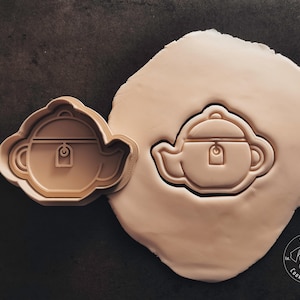 Könnte beinhalten: Ein brauner Plastik-Ausstecher in Form einer Teekanne mit einem Etikett an der Seite. Der Ausstecher ist auf einer hellbraunen Oberfläche mit einem weißen Abdruck der gleichen Teekannenform abgebildet.