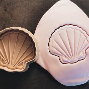 Könnte beinhalten: Eine Nahaufnahme eines rosa Silikon-Ausstechers in Form einer Muschel. Der Ausstecher liegt auf einer braunen Oberfläche. Der Ausstecher befindet sich neben einem beigen Plastik-Ausstecher in der gleichen Form. Der Plastik-Ausstecher hat ein detailliertes Design aus Linien und Kurven.