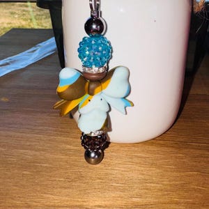 May include: A decorative keychain with a silver keyring. The keychain features a cluster of beads in silver, turquoise, and brown, along with a bow-shaped charm in shades of blue, yellow, and brown. The keychain is designed to be attached to keys or bags.