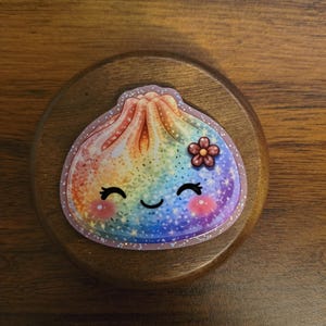 May include: A sticker shaped like a dumpling with a rainbow gradient finish and a glittery texture. The dumpling features a smiling face with closed eyes, pink cheeks, and a small brown flower accent on the side, all set against a clear border.