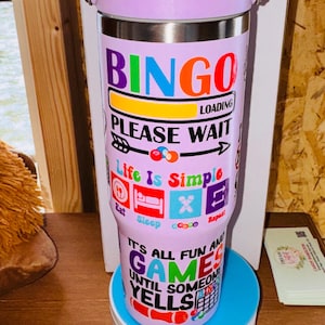 May include: A lavender and silver insulated tumbler with a handle. The tumbler has the word "BINGO" in colorful letters, along with the phrases "LOADING" and "PLEASE WAIT." Additional text includes "Life Is Simple" and "It's All Fun and Games Until Someone Yells."