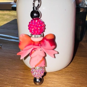 May include: A pink and orange handmade keychain with a bow and bead design. The keychain features a silver metal ring and a series of decorative beads in shades of pink and orange. The bow is a vibrant pink and orange color.