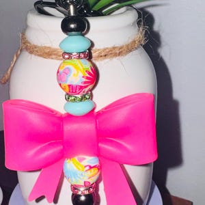 May include: A decorative keychain with a bright pink bow and colorful beads. The keychain has a silver ring and a black bead at the top and bottom of the beaded section. The beads have a floral design.