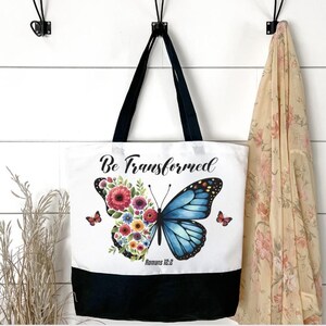 May include: White tote bag with black handles and a black bottom. The bag features a butterfly design with floral elements and the text "Be Transformed" and "Romans 12:2". Two small butterflies are on the sides of the main design.