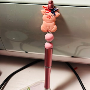 May include: A pink pen with a pink pig wearing a red, white, and blue bandana on its head. The pig is sitting on top of two pink beads.