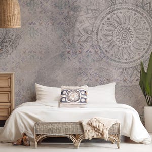 May include: A white bed with a white duvet cover and a white pillow. The bed has a white throw blanket at the foot of the bed and a small wooden bench in front of the bed. The wall behind the bed is a light gray with a large mandala design in gray and white. There is a small wooden dresser to the left of the bed and a potted plant to the right of the bed.