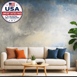 May include: A light-coloured sofa with two orange cushions and two dark blue cushions, positioned in front of a wall with a blue and grey watercolour-style mural. A wooden coffee table holds a small green plant and a dark bowl in the foreground.
