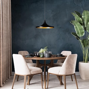 May include: A round wooden dining table with four upholstered beige chairs, featuring a black pendant light with a gold interior hanging above. The table is set with dark plates, glassware, and a small green plant in a glass vase.