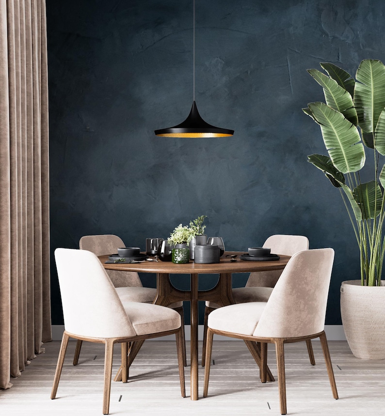 May include: A round wooden dining table with four upholstered beige chairs, featuring a black pendant light with a gold interior hanging above. The table is set with dark plates, glassware, and a small green plant in a glass vase.