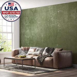 May include: A large wall mural featuring a distressed olive green texture with subtle vertical streaks and weathered patches. The mural is displayed behind a brown leather sofa with various patterned throw pillows and a wooden coffee table.