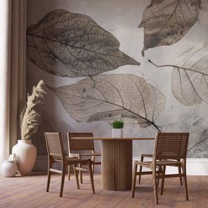 May include: A dining room set with a round wooden table and four woven-back chairs. The wall features a large-scale leaf design in shades of gray and brown. A vase with dried plants and a small potted plant are also visible.