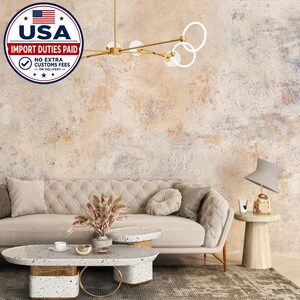 Könnte beinhalten: Ein getuftetes beiges Sofa mit passendem rundem Kissen, ein weißer Terrazzo-Couchtisch mit goldenem Ringfuß, ein goldener Kronleuchter mit kreisförmigen Akzenten und eine graue Tischlampe auf einem zylindrischen Beistelltisch vor einer strukturierten Wand.