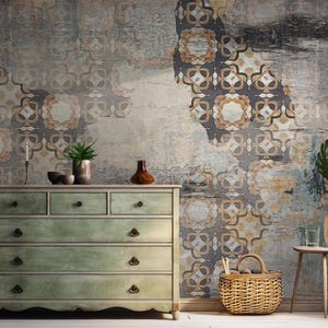 May include: A light green dresser with six drawers and ornate brass handles sits in front of a wall with a distressed, patterned wallpaper. The wallpaper features a geometric design in shades of brown, gray, and beige. A wicker basket sits on the floor in front of the dresser.